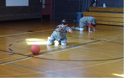 As student wearing camoflauge military gear and a blind-fold kneels in a basketball court with her arms outstreched searching for the ball.