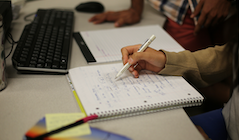 A student writes in their notebook with a computer in the background.