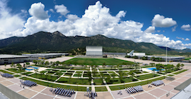 A landscape photo of a campus buildings with a blue sky, mountains, and grass.